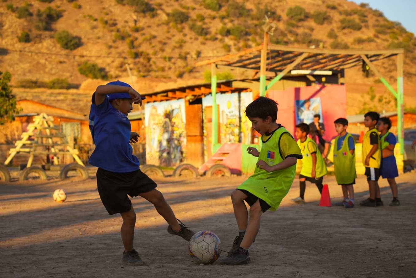 Dos niños disputan el balón en la cancha de tierra, con el cerro y las casas del barrio al fondo