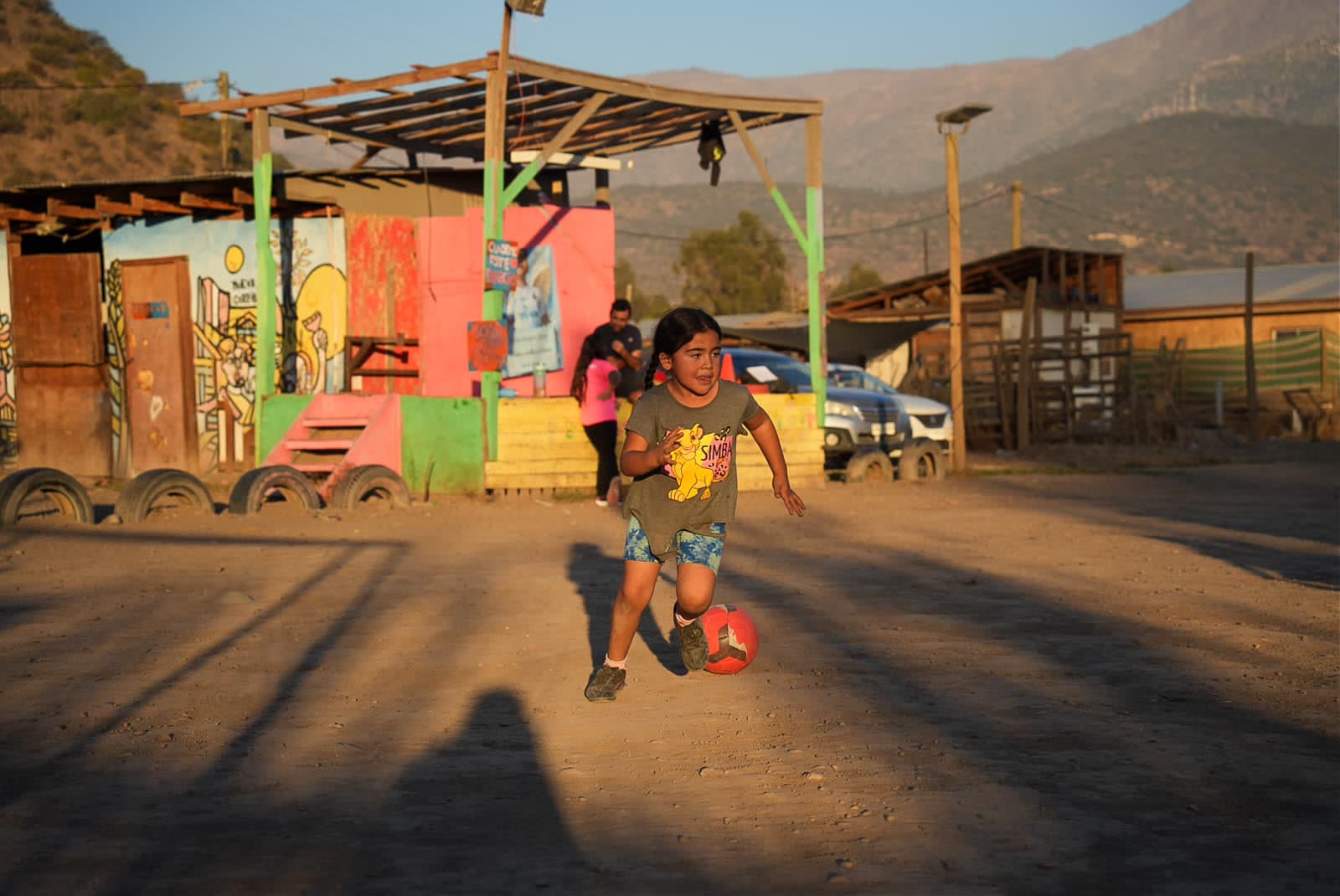 Una niña corre con el balón en la cancha de tierra del barrio, frente a un muro pintado de colores, al atardecer