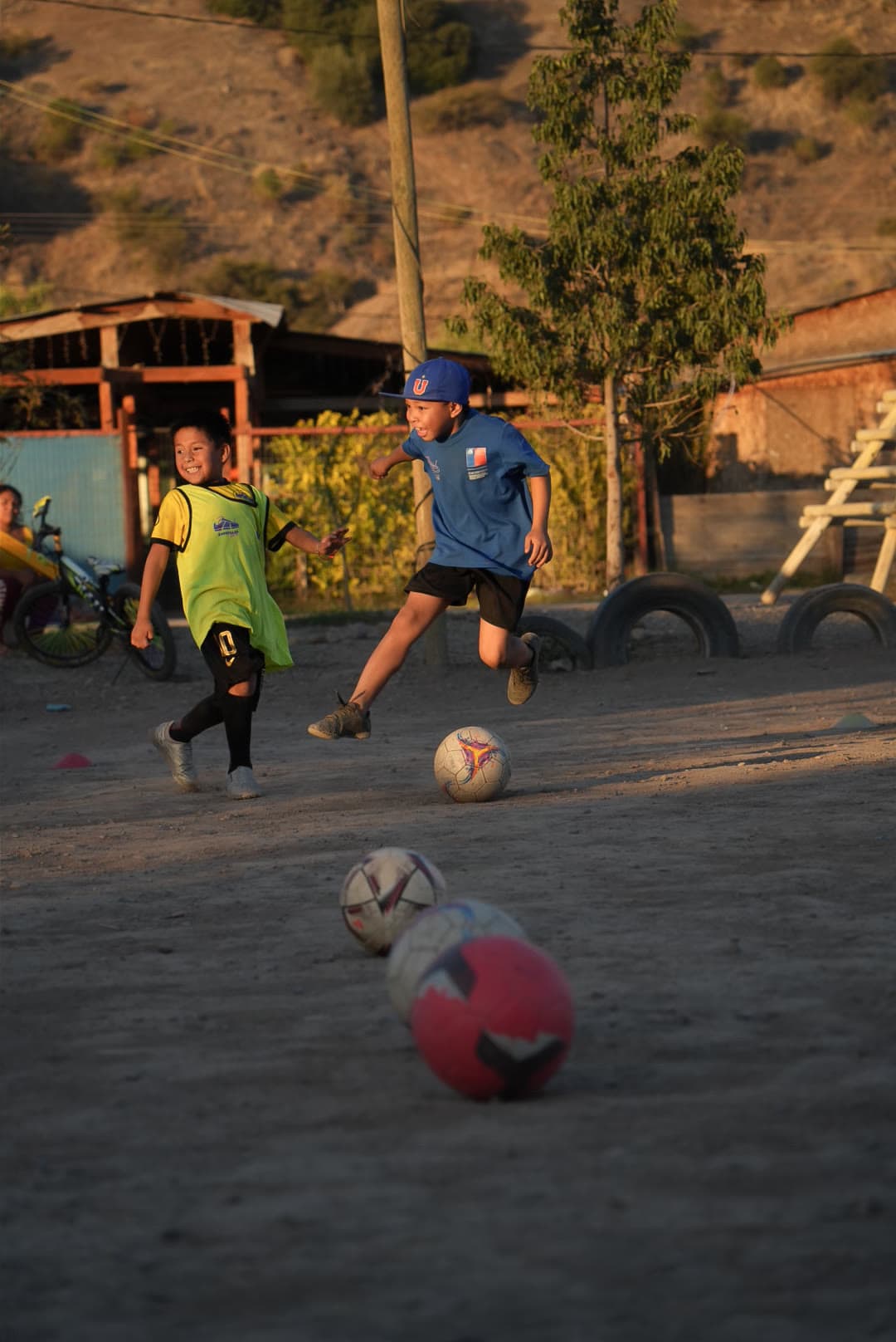 Niño remata el balón en la cancha de tierra, con el cerro y el barrio de fondo